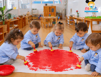 Niños y niñas de Educación Infantil de los Centros Escolares Menuts, vestidos con sus baberos de rayas azules y blancas con el logotipo del centro, pintan juntos un gran círculo rojo en el suelo con pinceles infantiles en un aula Montessori.