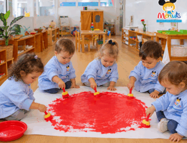 Niños y niñas de Educación Infantil de los Centros Escolares Menuts, vestidos con sus baberos de rayas azules y blancas con el logotipo del centro, pintan juntos un gran círculo rojo en el suelo con pinceles infantiles en un aula Montessori.