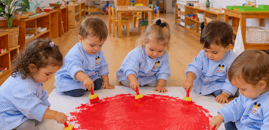 Niños y niñas de Educación Infantil de los Centros Escolares Menuts, vestidos con sus baberos de rayas azules y blancas con el logotipo del centro, pintan juntos un gran círculo rojo en el suelo con pinceles infantiles en un aula Montessori.